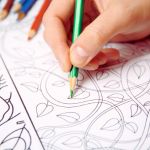 A hand colours an intricate floral pattern with a green pencil. Other coloured pencils lie nearby. The scene conveys focus and creativity.