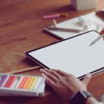 A person uses a stylus on a blank tablet screen on a wooden desk. Nearby are coloured pencils and papers, suggesting a creative and focused atmosphere.